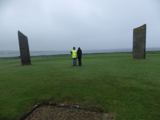 Two young men in coats and hats on a misty, gray day, standing between two large (approximately 3 meters tall) standing stones at a neolithic site in Orkney. 